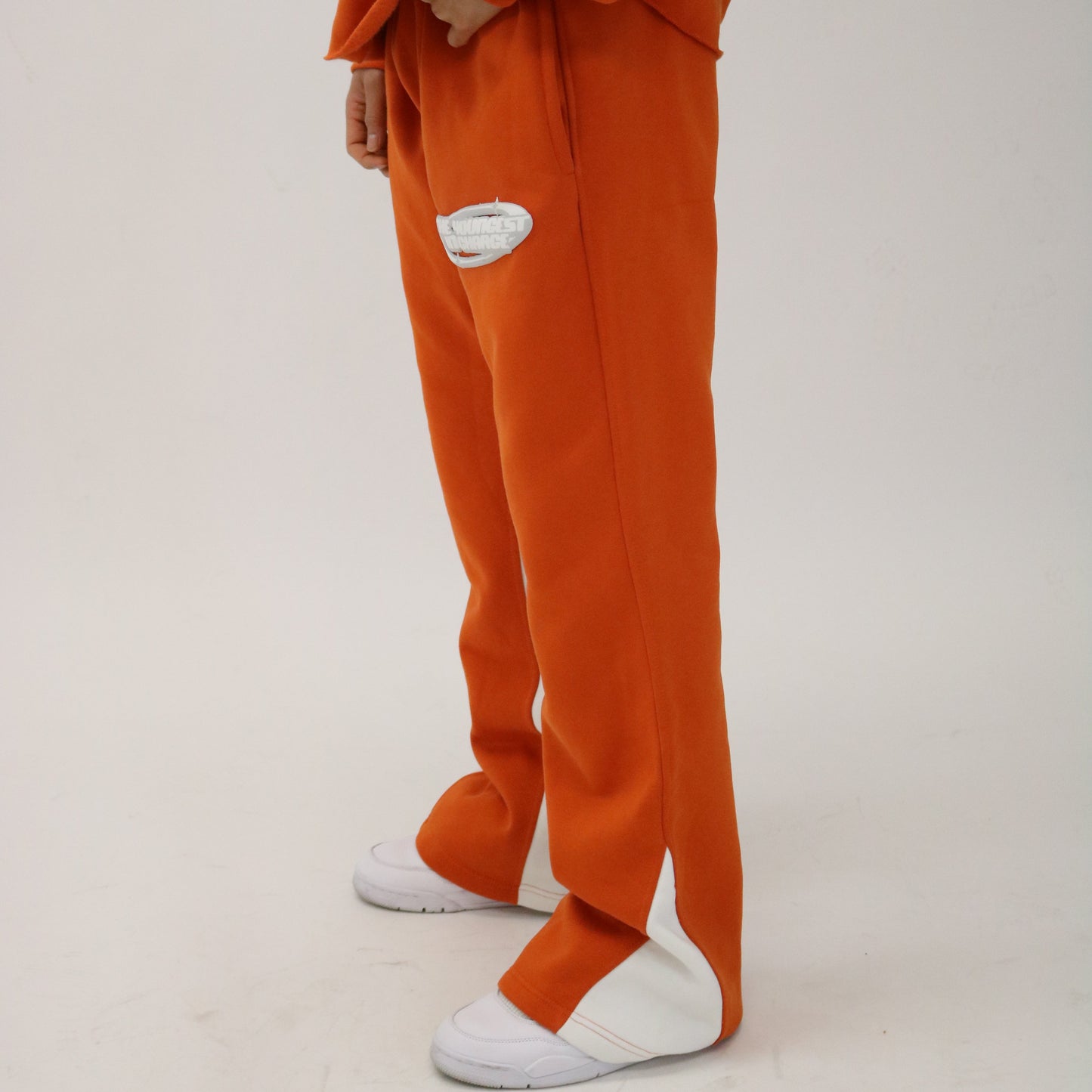 A person wearing orange stacked pants with a puff print logo on the chest, back, and jogger. They are also wearing a matching orange top, white sneakers, and the photo is taken from a side angle.