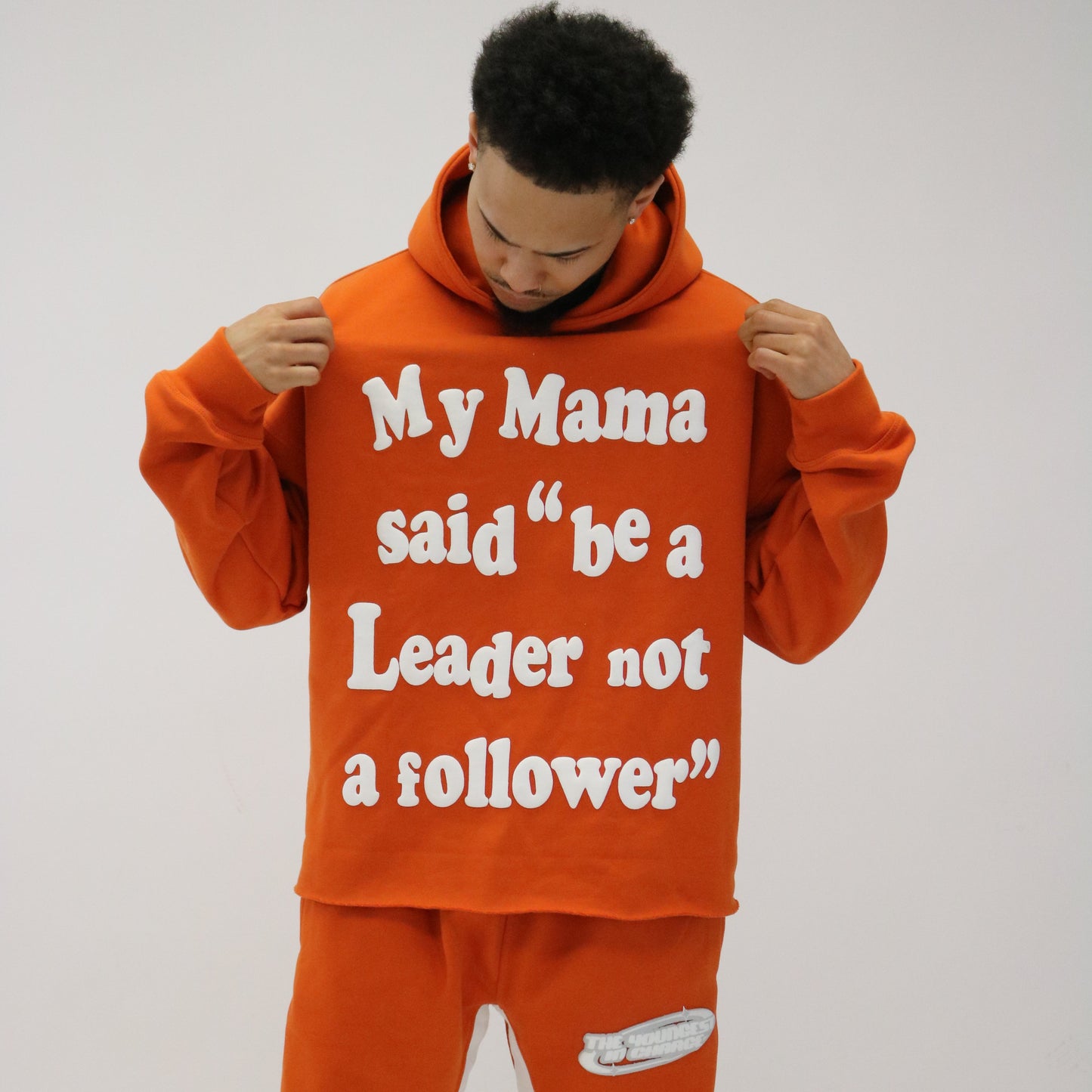 A person wearing an orange hoodie with the text 'My Mama said be a Leader not a follower' printed on it, paired with matching orange tracksuit bottoms, standing against a neutral background.
