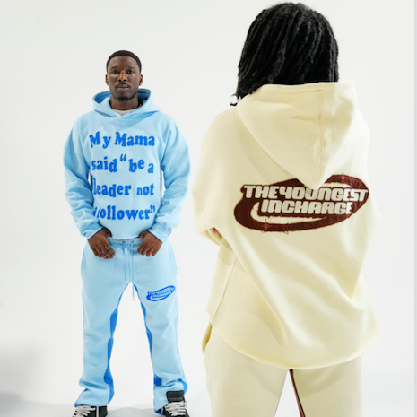 Two models wearing matching sweatshirt and jogger sets on a white studio background. One model faces forward wearing a light blue tracksuit with blue text on the chest, and the other model is shown from the back wearing a cream-colored tracksuit with a brown graphic on the back reading 'THE YOUNGEST UNCHANGED'.